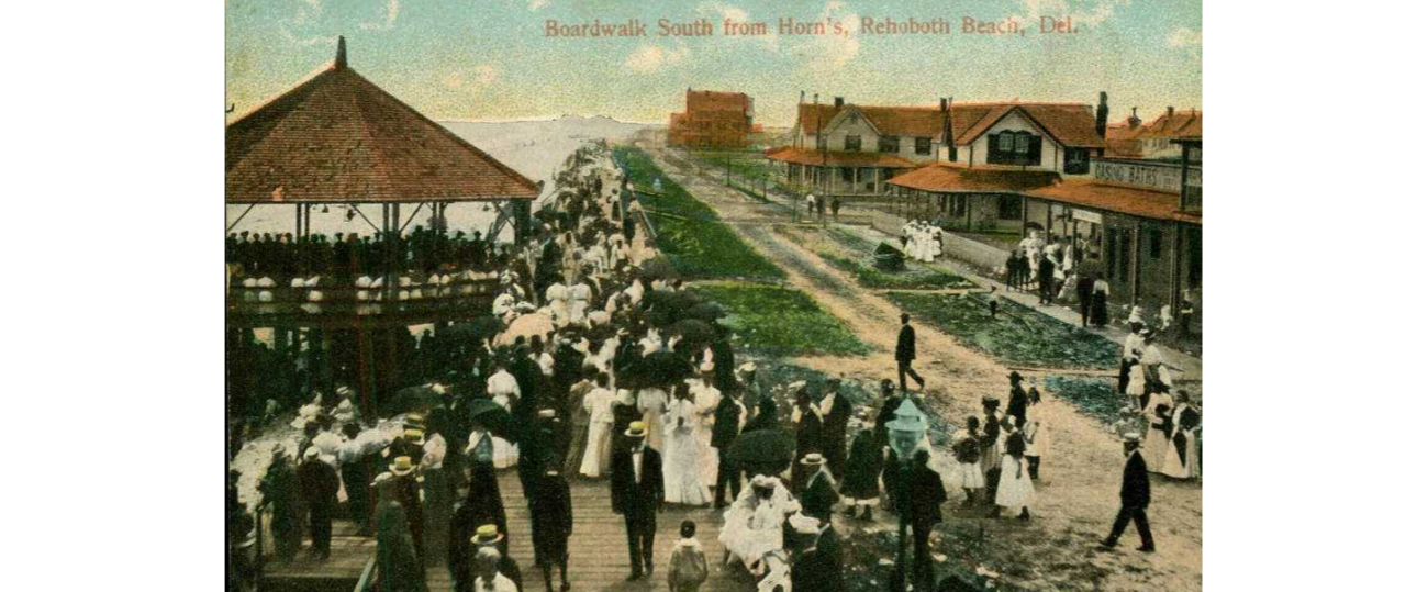 A historic photo, depicting men in suits and women in long dresses, walking along the boardwalk in Rehoboth Beach