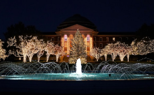 Nighttime. A very large university building in the background, with several trees, decorated with christmas lights, and a fountain in the front.