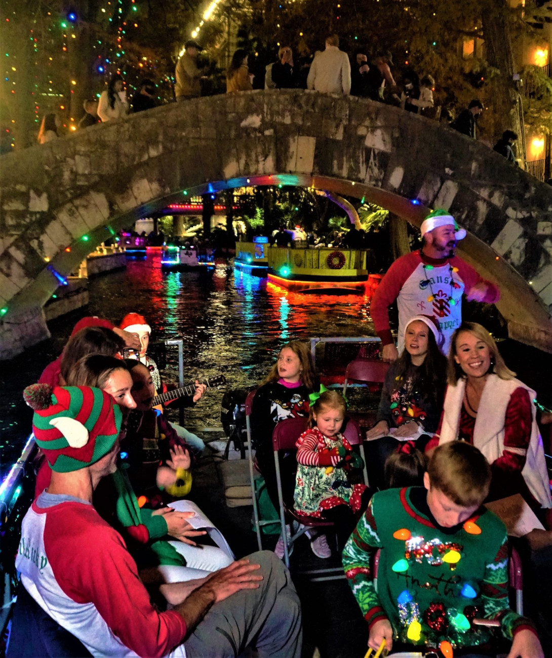People, on a boat, cruising slowly down a river, singing carols.
