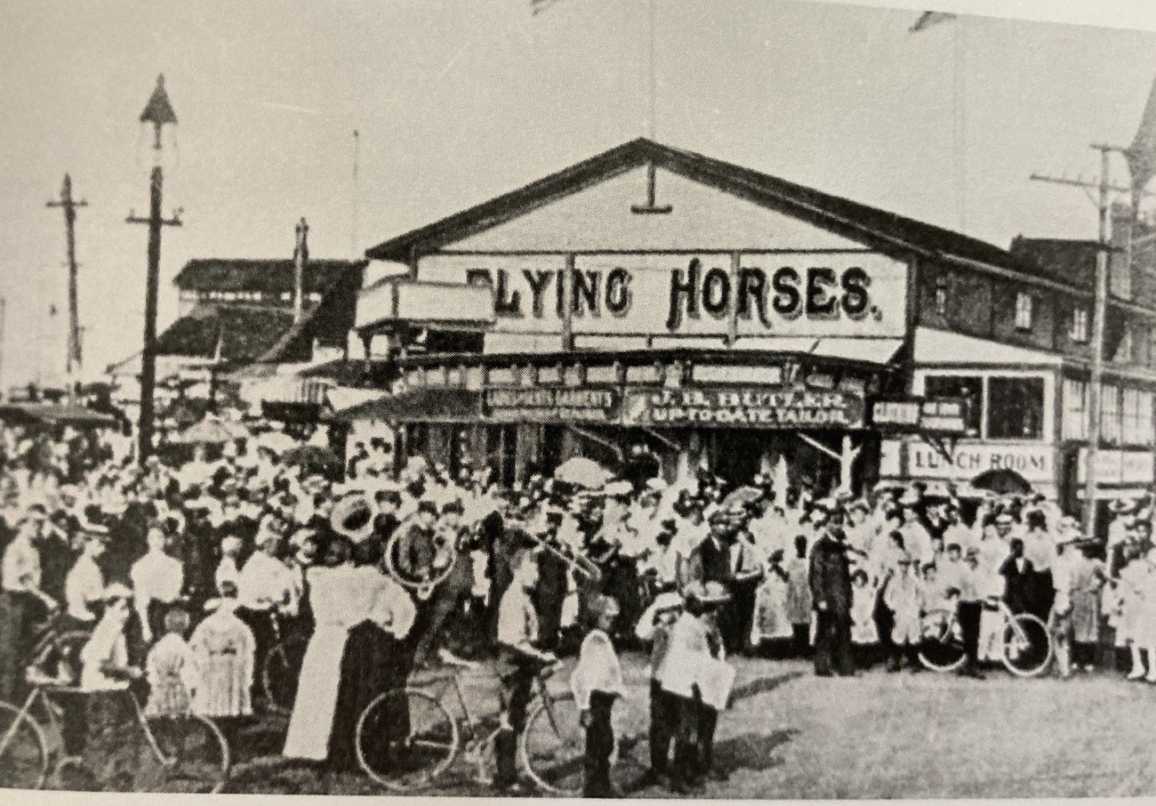 A black and white photo, historic.  A crowd of people, some with bicycles, stand in front of a one-story building with the sign "Flying Horses" on it.
