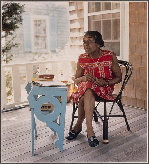 A woman sits outside her home. She has a blue desk in front of her.