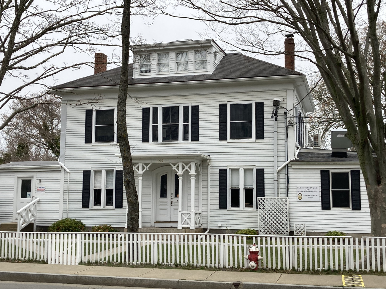 A white two-story building, with an attic, stands.  Black shutters.  White picket fence in front.
