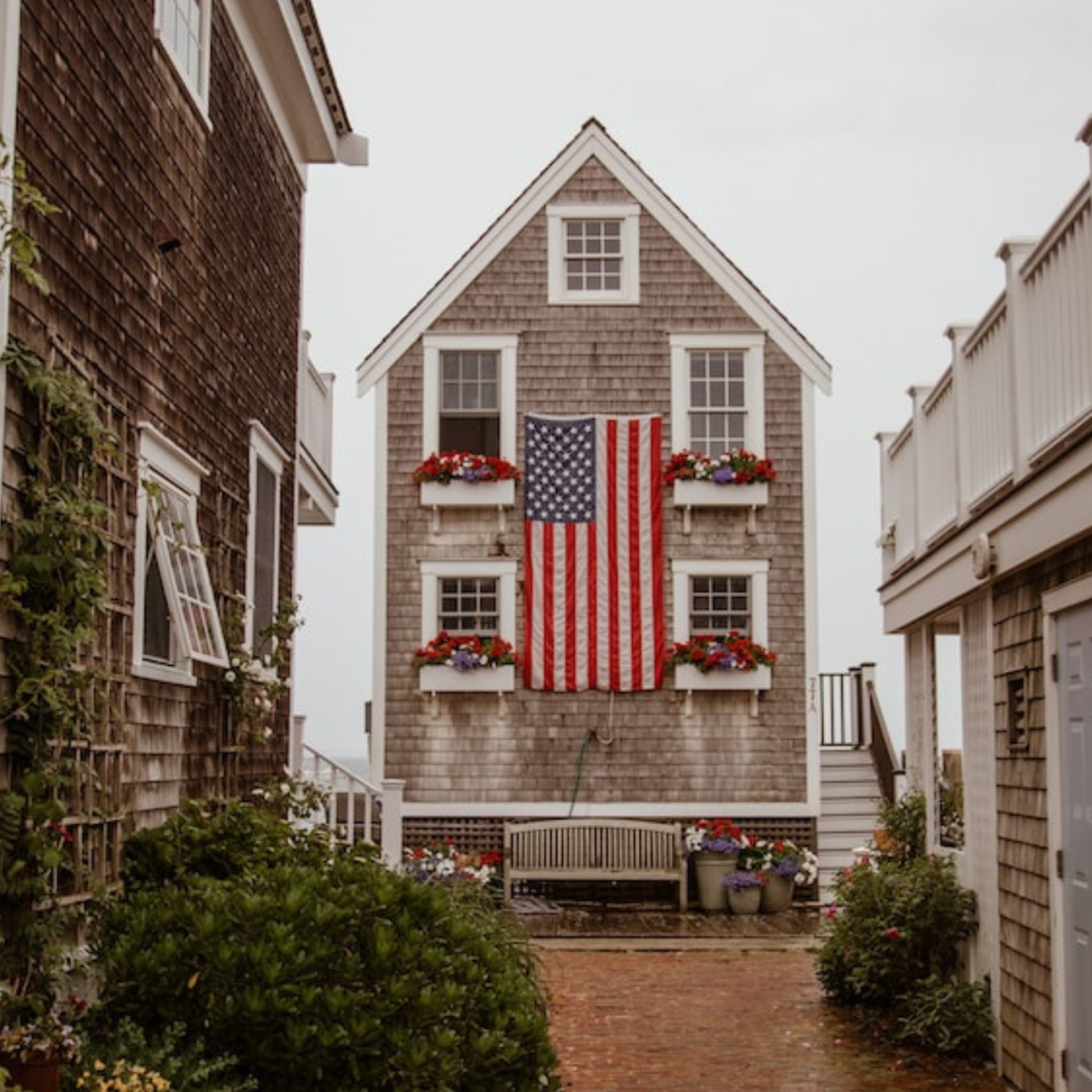 a pretty, brown shingled house, with an American flag hung in the center. red and white flowers in the flower boxes at each window.