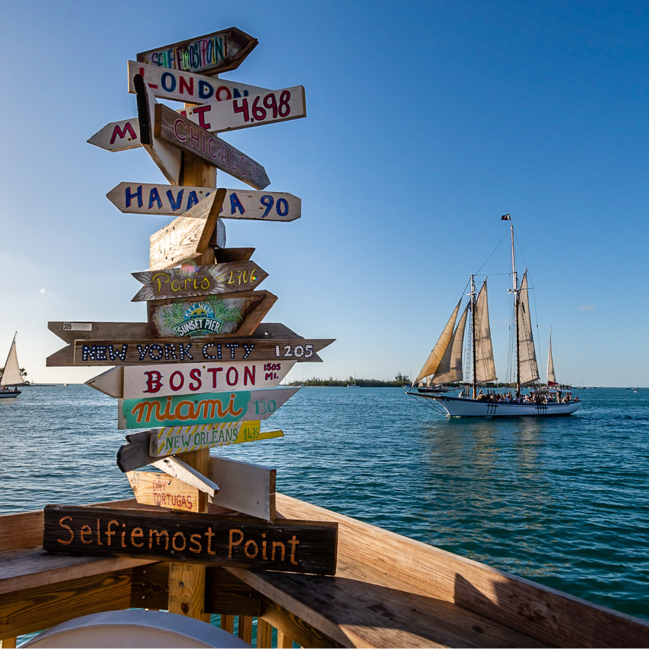 Signs at the southernmost point of Key West, and the USA. "Selfiemost Point", "Boston", "Miami" are posted. A sailboat on the ocean is in the background.