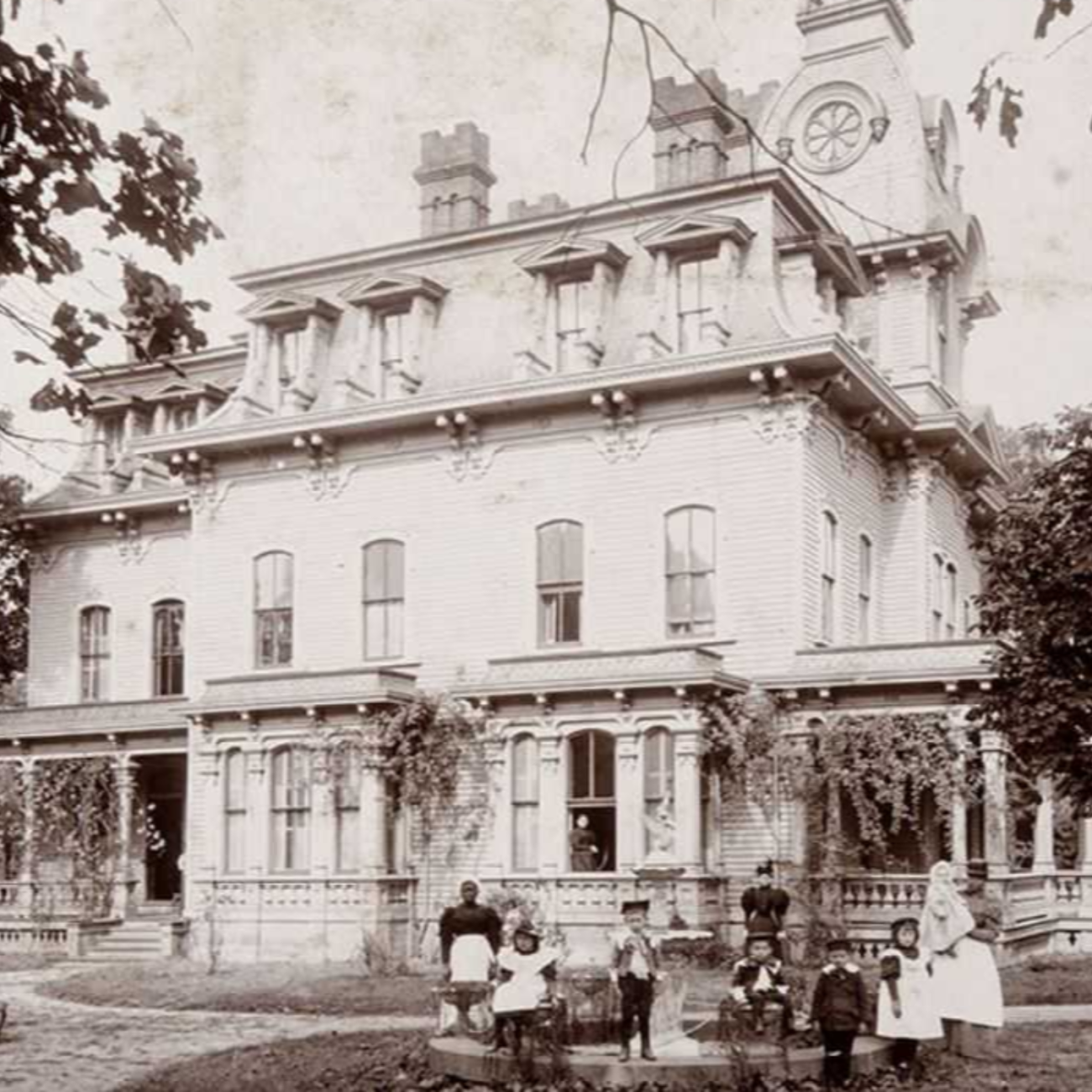 a black and white historic photo of a mansion, with a family sitting in the front lawn.