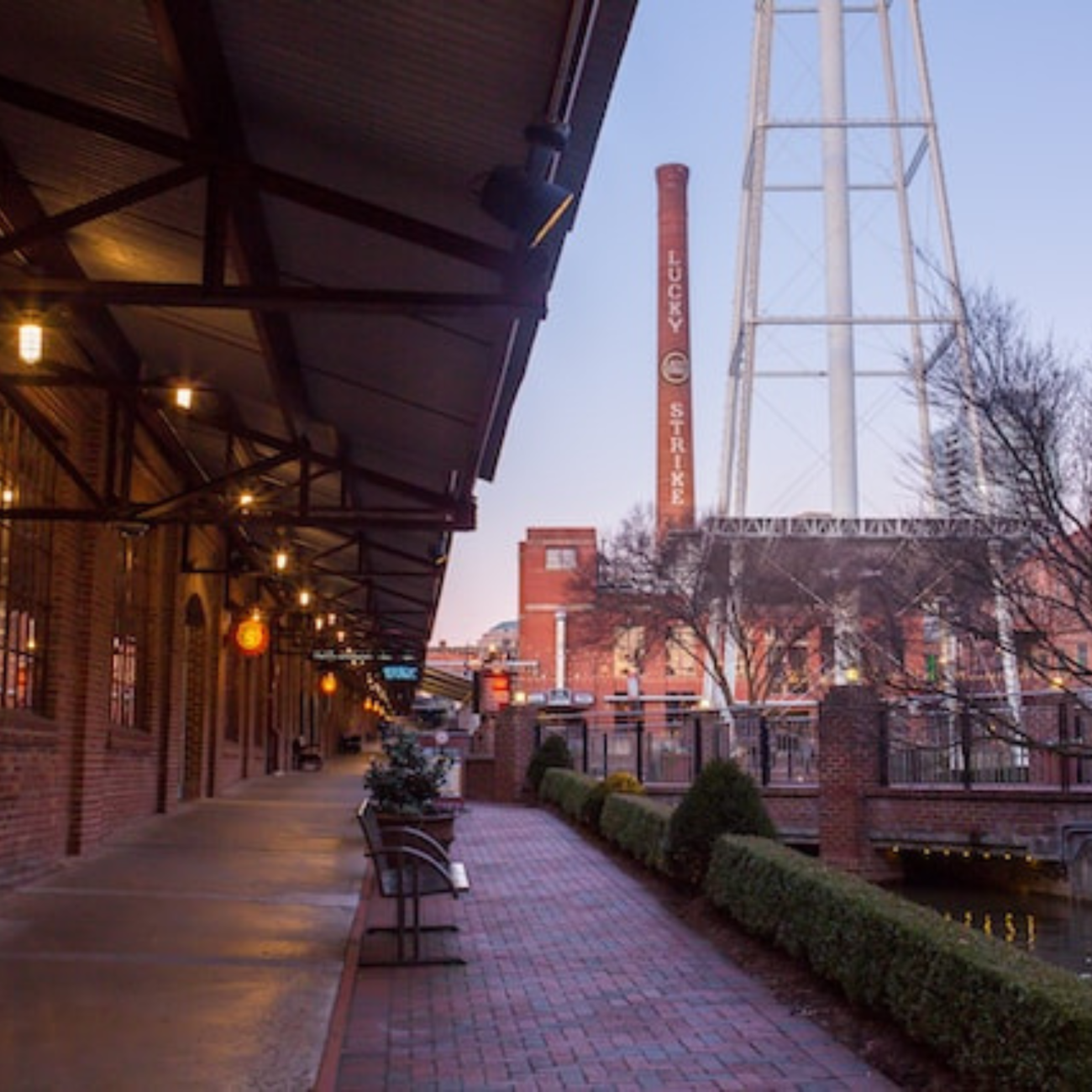 a brick sidewalk, next to a flowing waterway, in a busy urban center. A brick tower in background, next to a water tower.