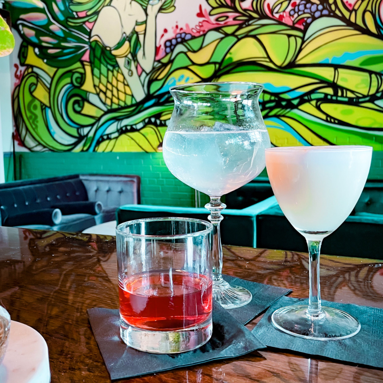 Three cocktails on a table, a colorful green mural in background.
