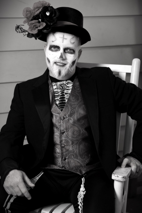 A black & white photo of a man, or spirit, seated in a rocking chair. He wears a three-piece suit, and a top hat.