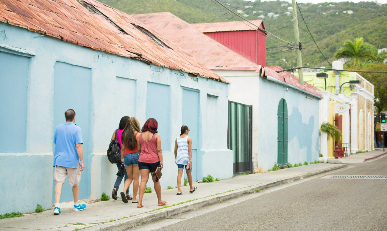Five tourists walk along the sidewalk; a light blue brightly colored buildings beside them, a mountain dotted with homes in the background.