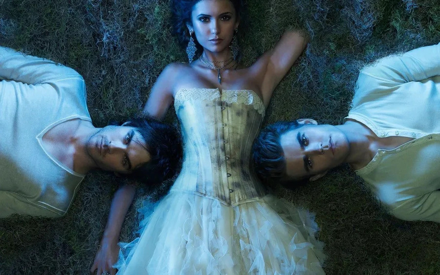 Two men, from the Vampire Diaries universe, lay on either side of a woman. She is wearing a white driess.